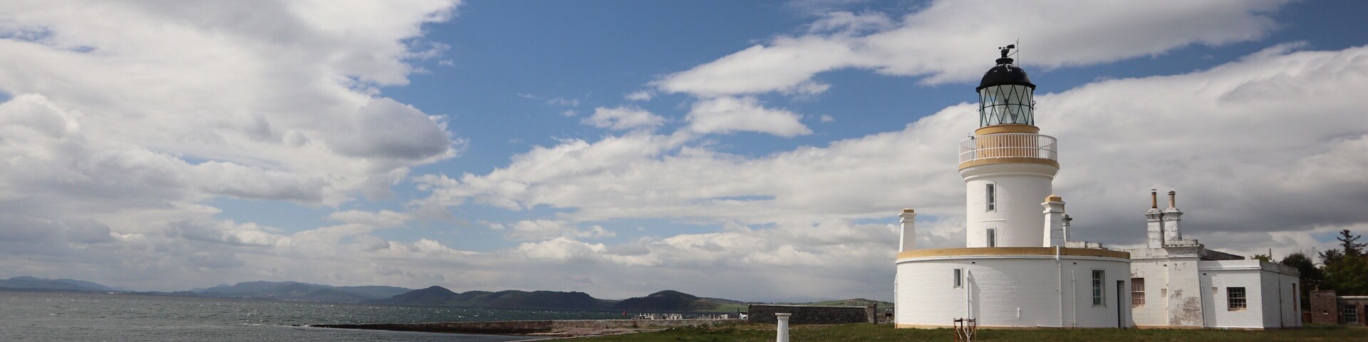 Un faro con exterior blanco y parte superior verde, ubicado en una colina cubierta de hierba con vistas a una masa de agua y montañas al fondo.