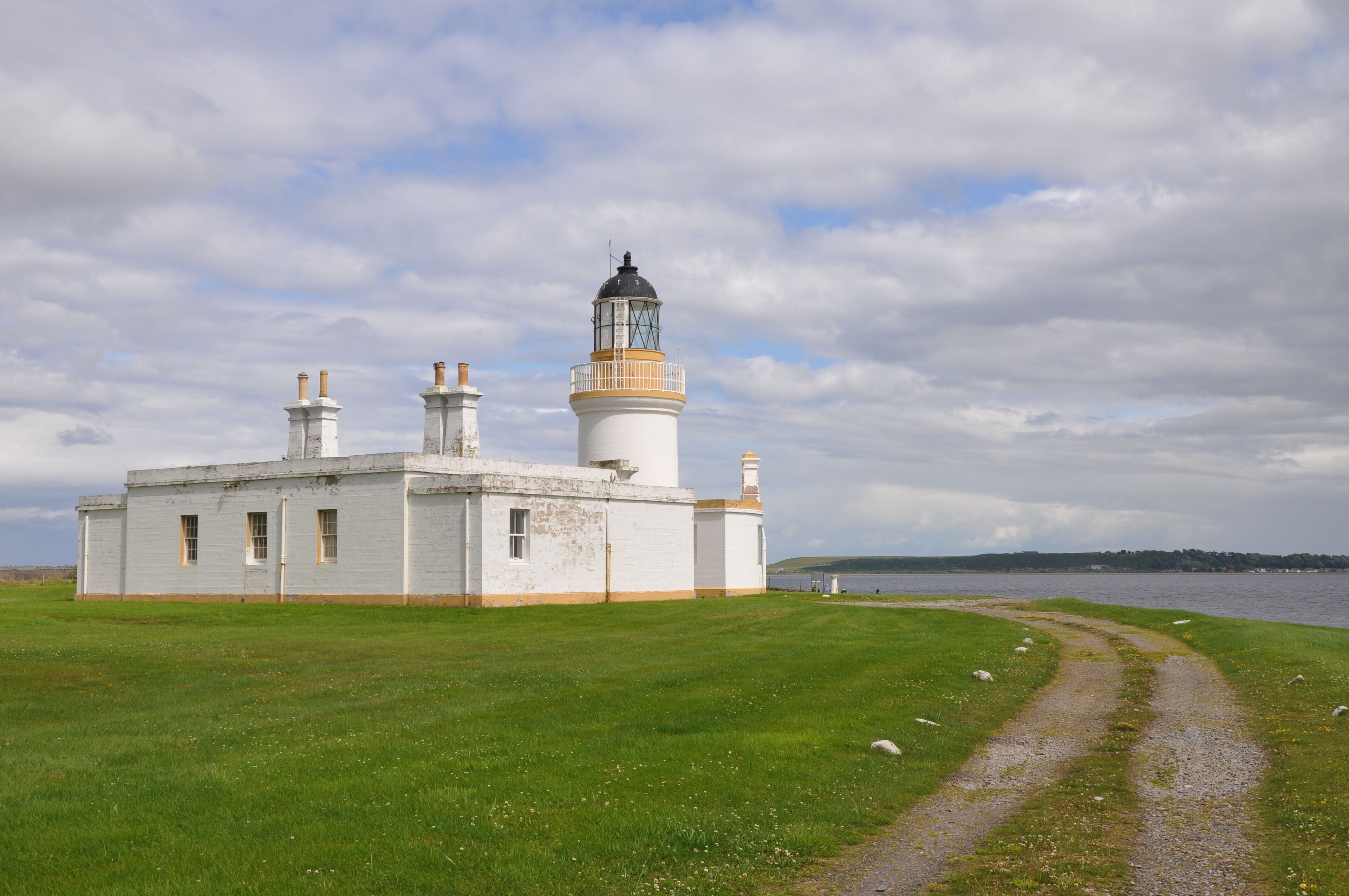 A lighthouse with a white and yellow exterior, a grassy field, and a body of water in the background.