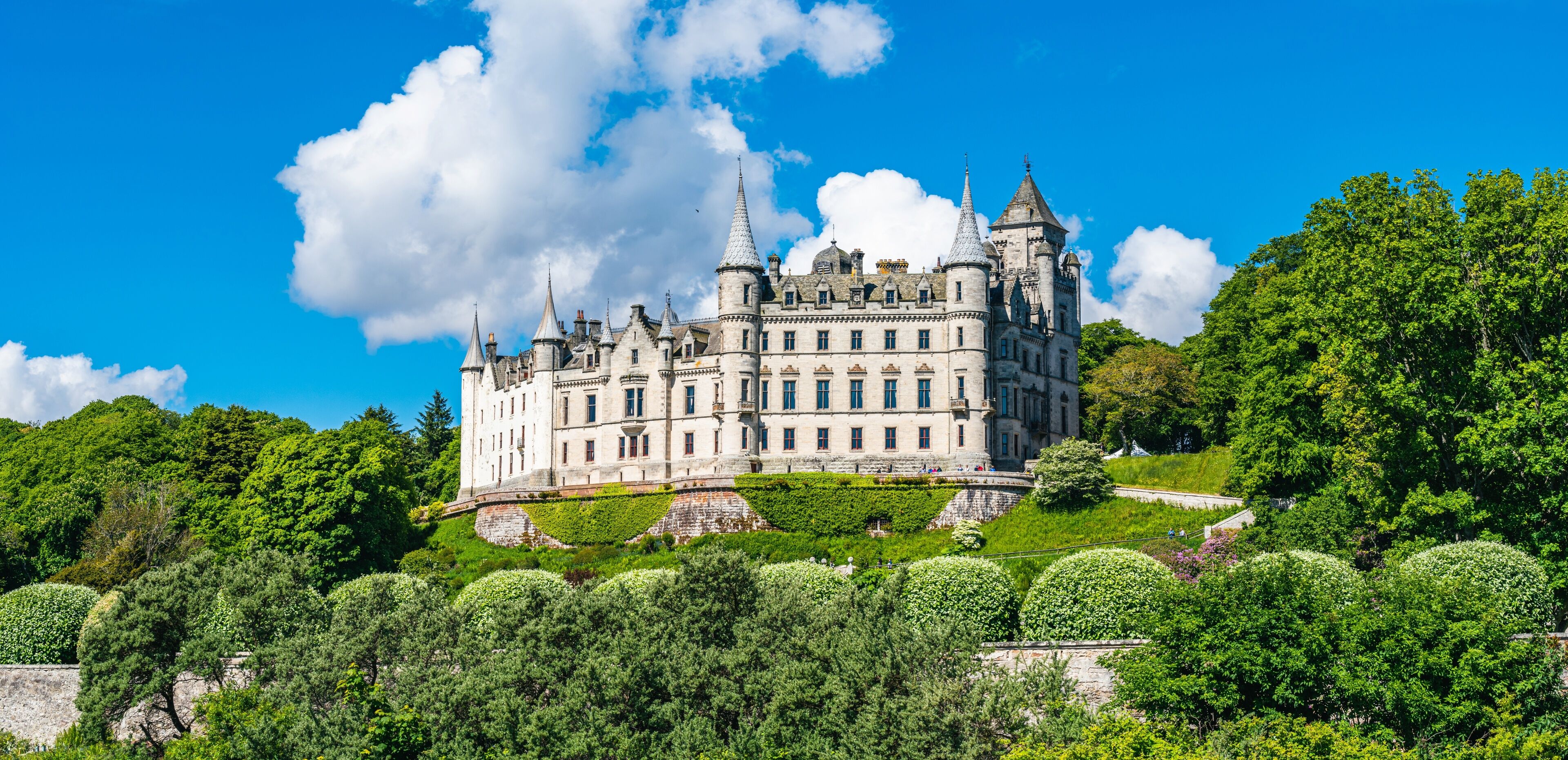 A large, historic castle surrounded by lush greenery and a clear blue sky.