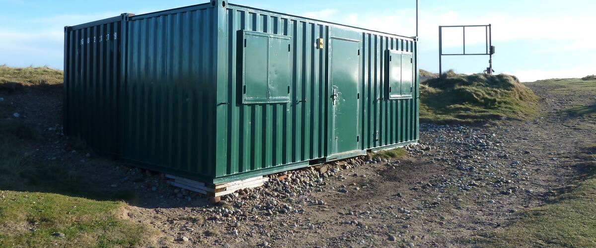 A green metal container with the number 6U2179 on it, situated on a gravel path with grass and hills in the background.