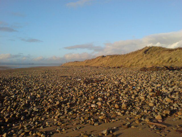 Shore at Kenfig nature Reserve Kenfig Nature Reserve is a large area of sand dunes and buroughs. The three dark structures visible in the distance are cranes, part of Margam steel works. On the map, this area is described as Kenfig Sands - those are pretty big sand grains though...