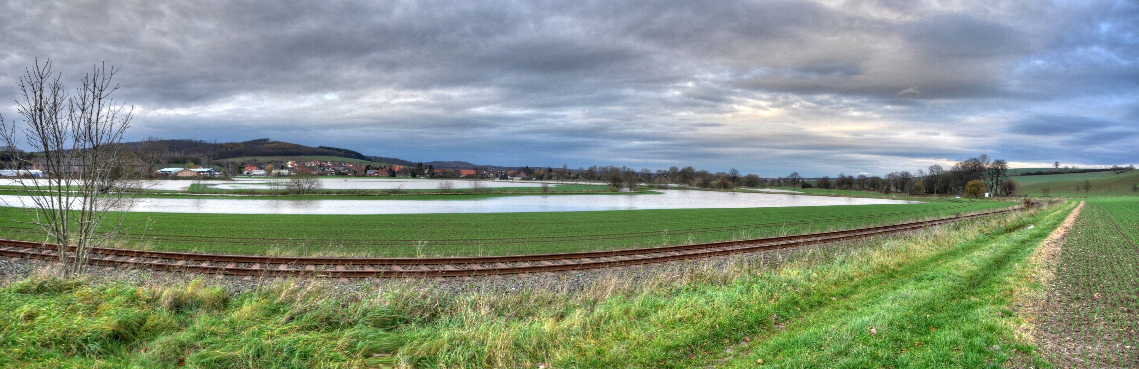 A rural landscape with a large pond, green fields, a railway track, and distant houses.