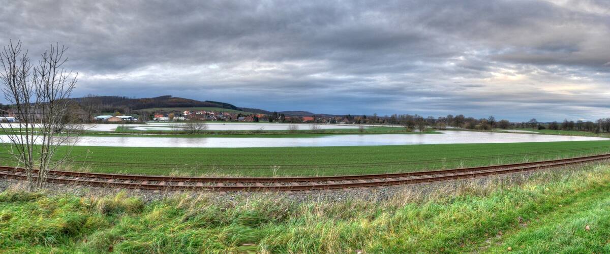 A rural landscape with a large pond, green fields, a railway track, and distant houses.