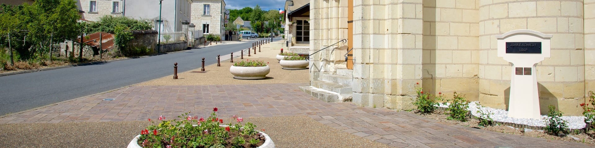 A street with a stone building, a large flower bed, and a sign reading "John F. Kennedy Loop".