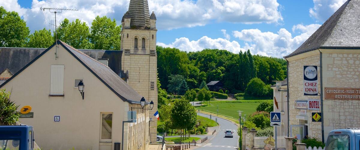 A quaint village street with a church tower, a stone building with a sign for 'Chez Dina', and a blue van parked on the side.