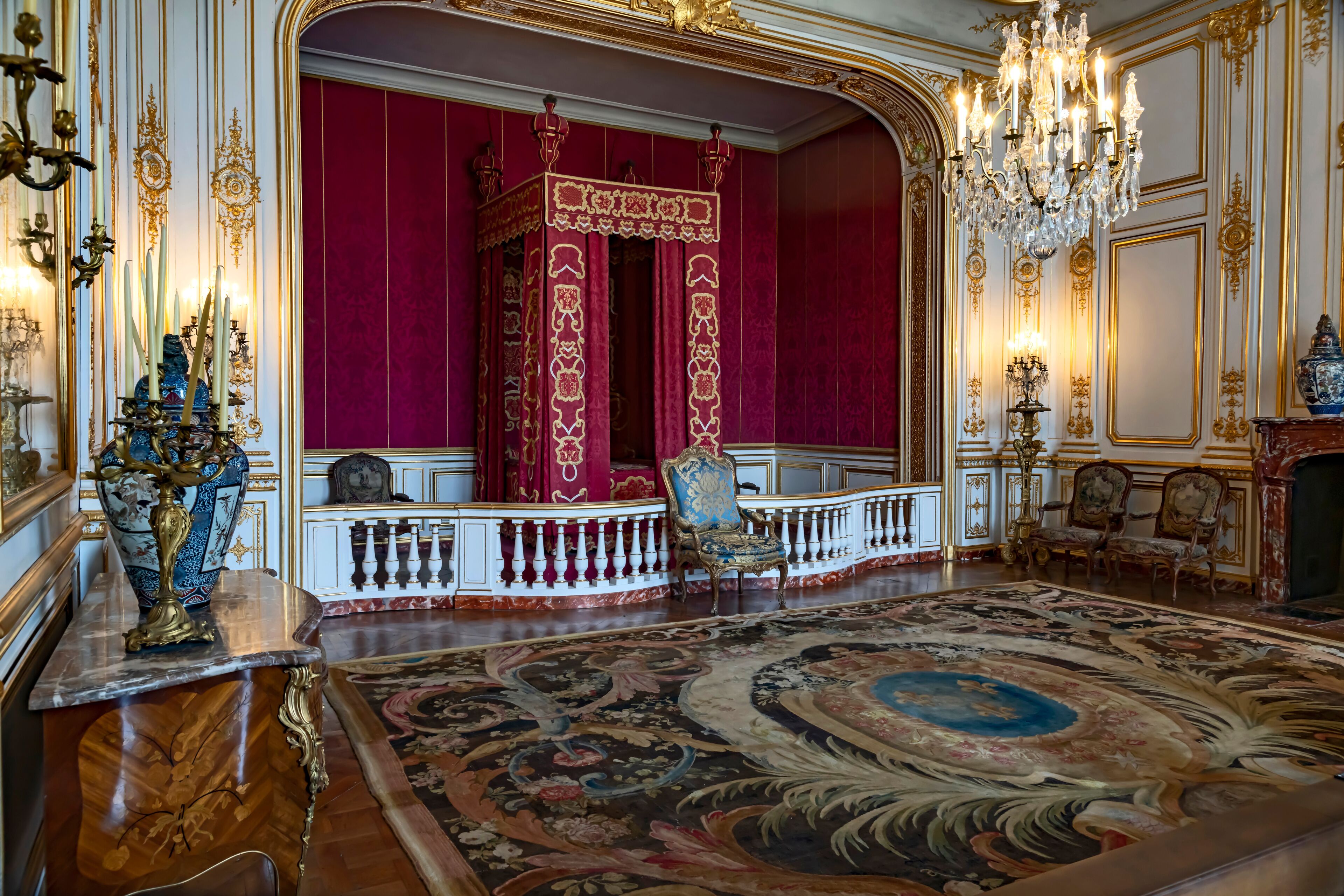 An opulent room with a large ornate carpet, a red velvet backdrop, and a chandelier.