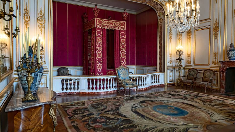 An opulent room with a large ornate carpet, a red velvet backdrop, and a chandelier.