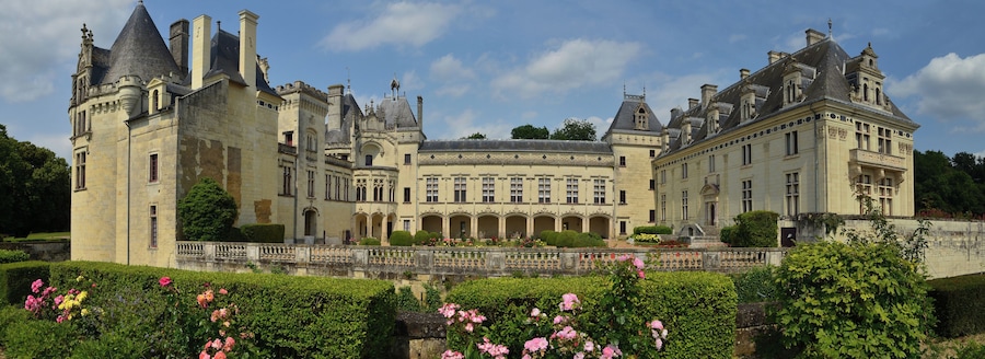 Ein großes, historisches Schloss mit zahlreichen Türmen und einem gepflegten Garten mit rosa Blumen.