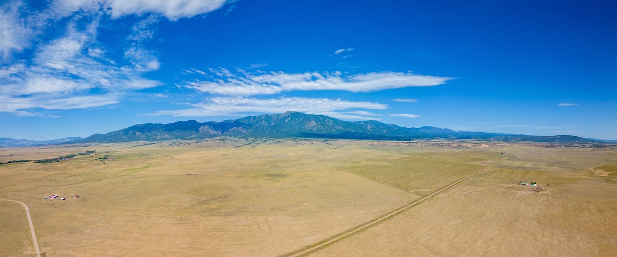 A vast open field with scattered buildings and a mountain range in the distance under a clear blue sky.