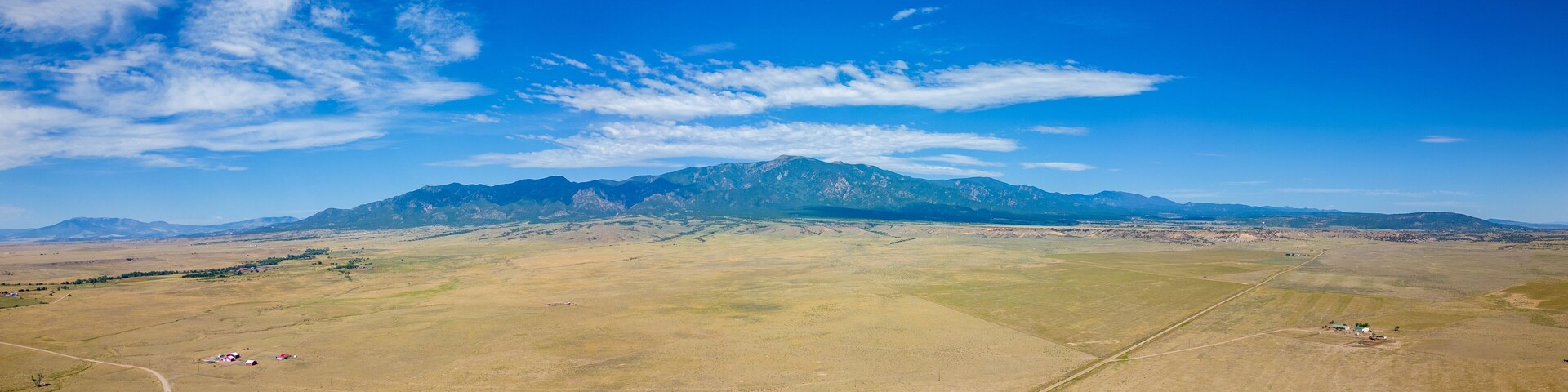 A vast open field with scattered buildings and a mountain range in the distance under a clear blue sky.