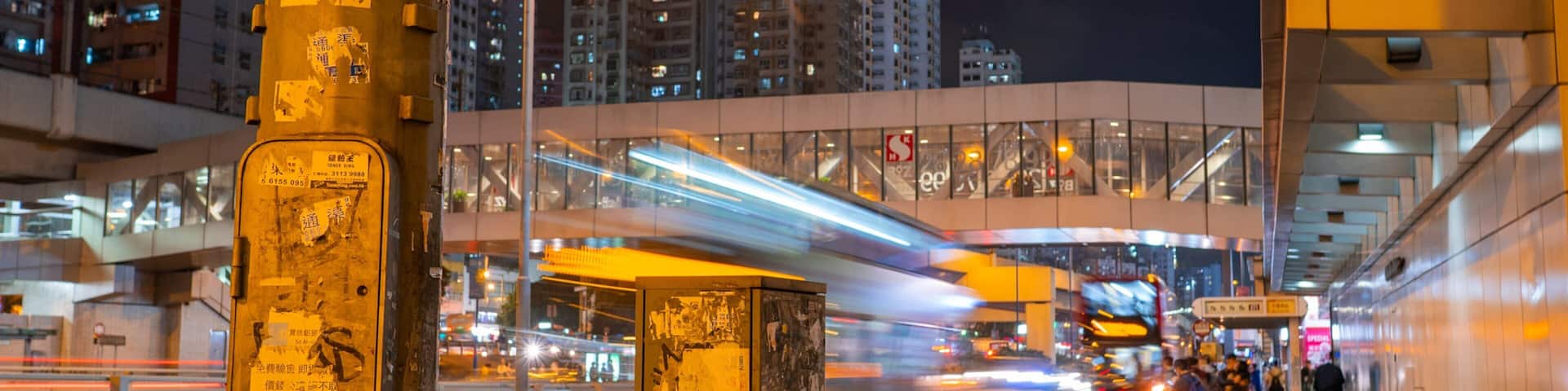 A nighttime city street with a moving train, tall buildings, and graffiti on a pole.