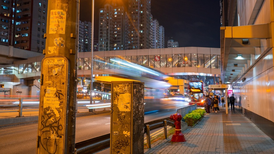 A nighttime city street with a moving train, tall buildings, and graffiti on a pole.