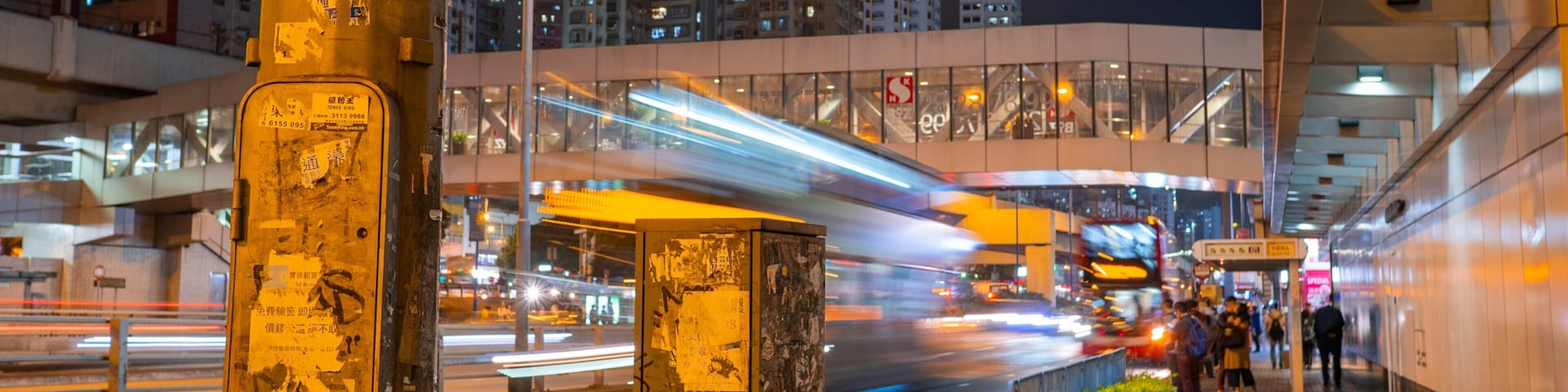 A nighttime city street with a moving train, tall buildings, and graffiti on a pole.