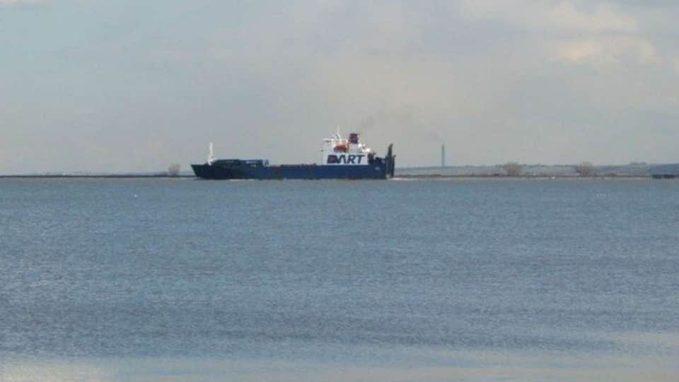A large cargo ship on calm waters with a distant shoreline and a smokestack in the background.