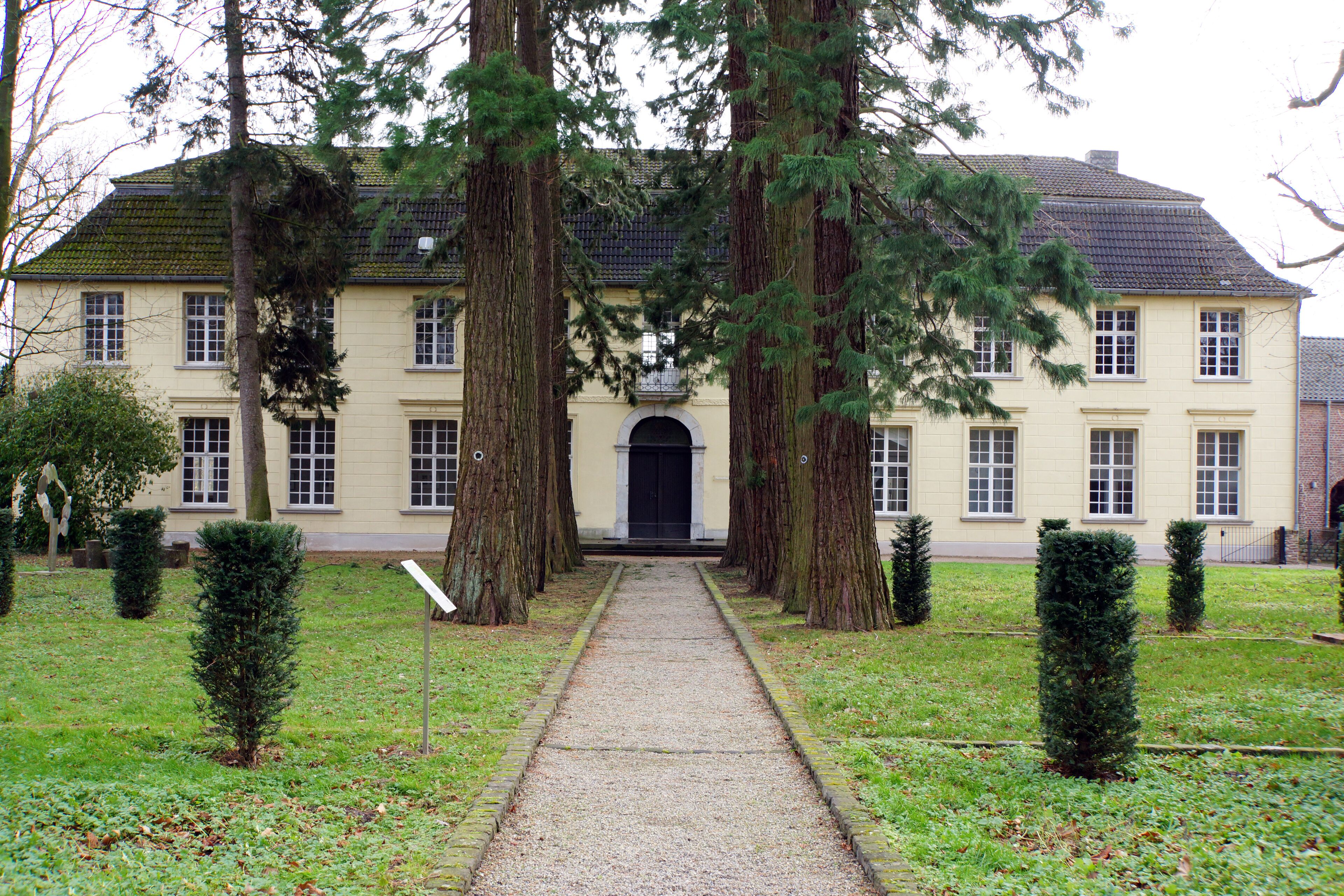 A large, cream-colored building with a symmetrical facade, surrounded by a gravel path and several tall, slender trees.