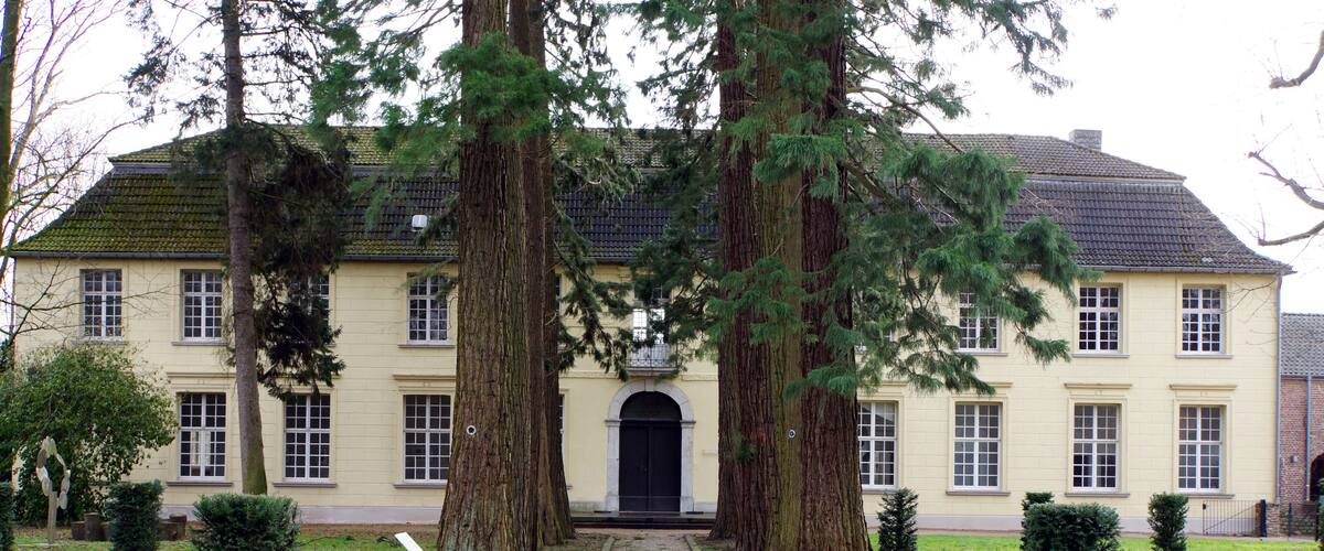 A large, cream-colored building with a symmetrical facade, surrounded by a gravel path and several tall, slender trees.