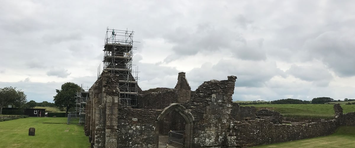 A historic stone structure with scaffolding, surrounded by a grassy field and trees.
