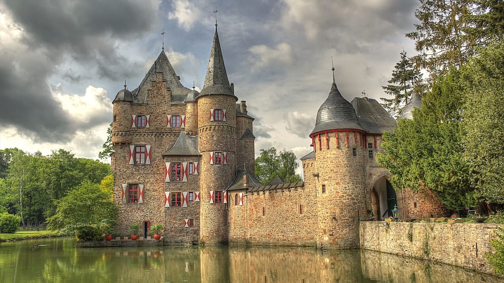 A historic stone castle with multiple towers, a moat, and red window shutters.