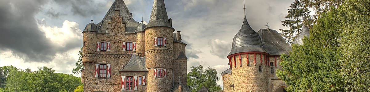 A historic stone castle with multiple towers, a moat, and red window shutters.