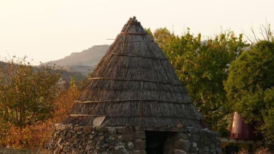 A traditional thatched-roof hut with a stone base, surrounded by greenery and trees.
