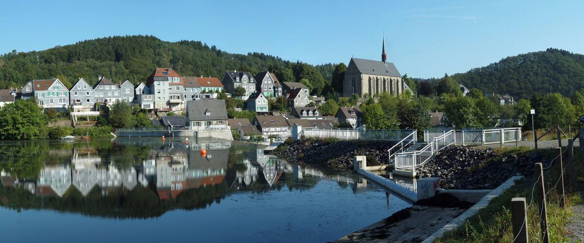 A calm river reflecting buildings, a church, and a hill with dense forest.