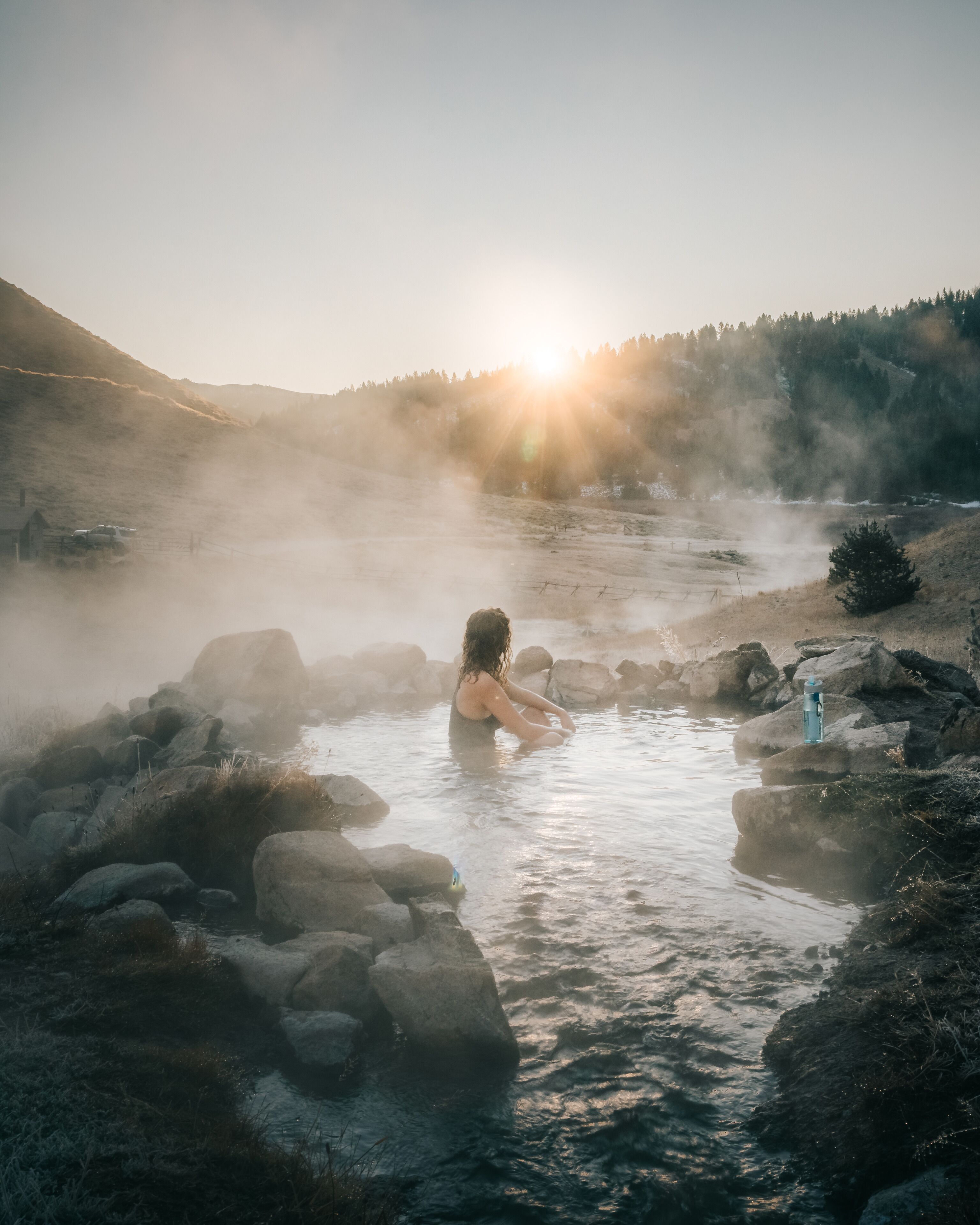 A person in a natural hot spring surrounded by rocks and trees with the sun setting in the background.
