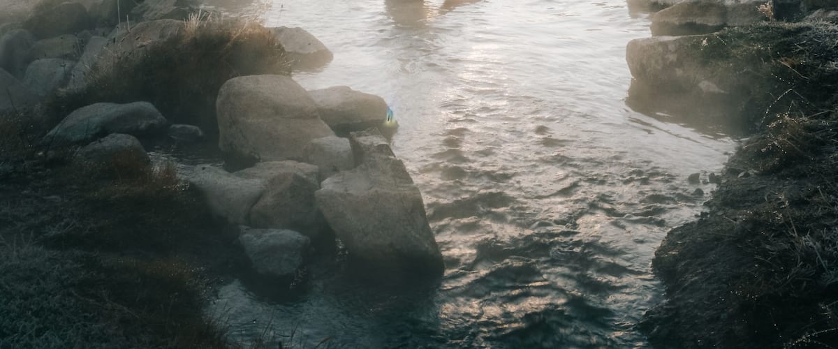 A person in a natural hot spring surrounded by rocks and trees with the sun setting in the background.