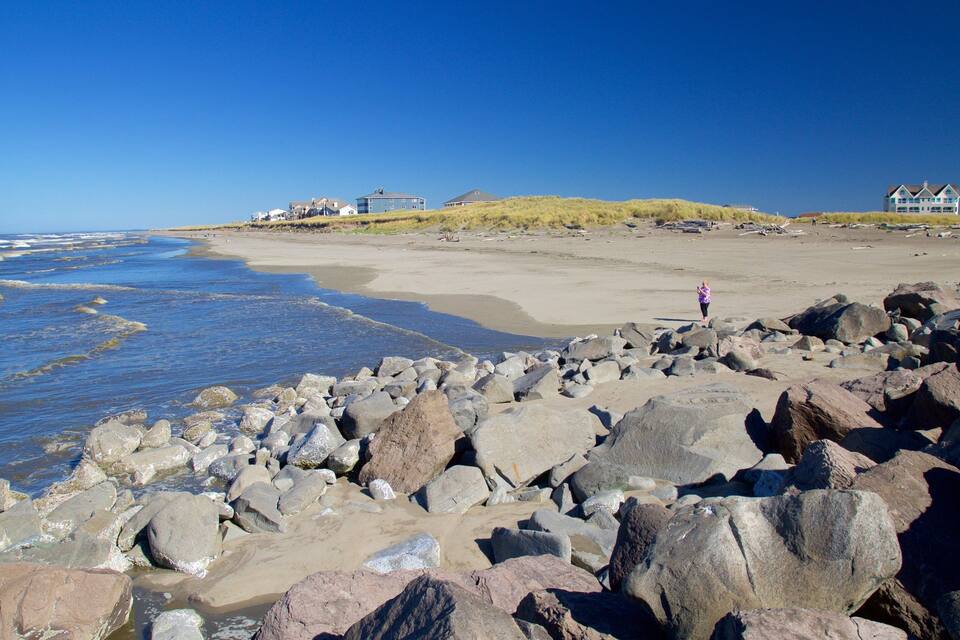 Ein felsiger Strand mit großen Steinen, ein sandiger Uferbereich und Häuser im Hintergrund.