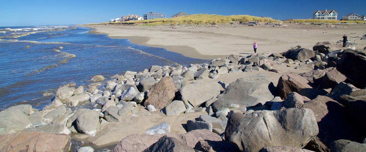 A rocky beach with large boulders, a sandy shore, and houses in the background.