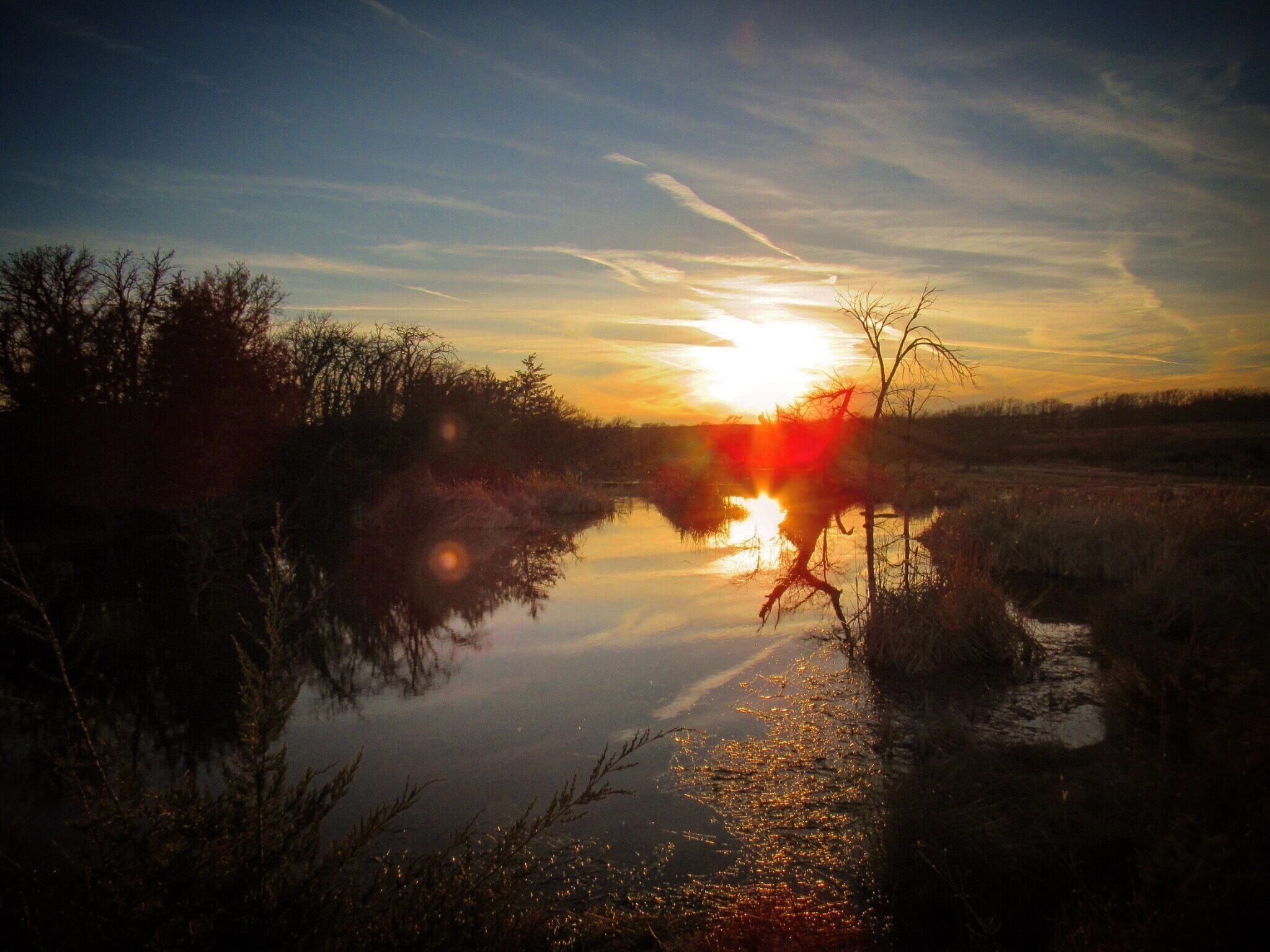 A sunset over a calm river with reflections of the sky, surrounded by trees and shrubs.