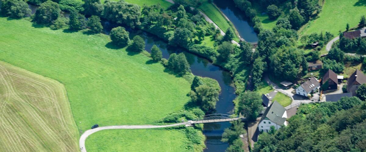 An aerial view of a river winding through a green landscape with fields and a bridge connecting two areas.