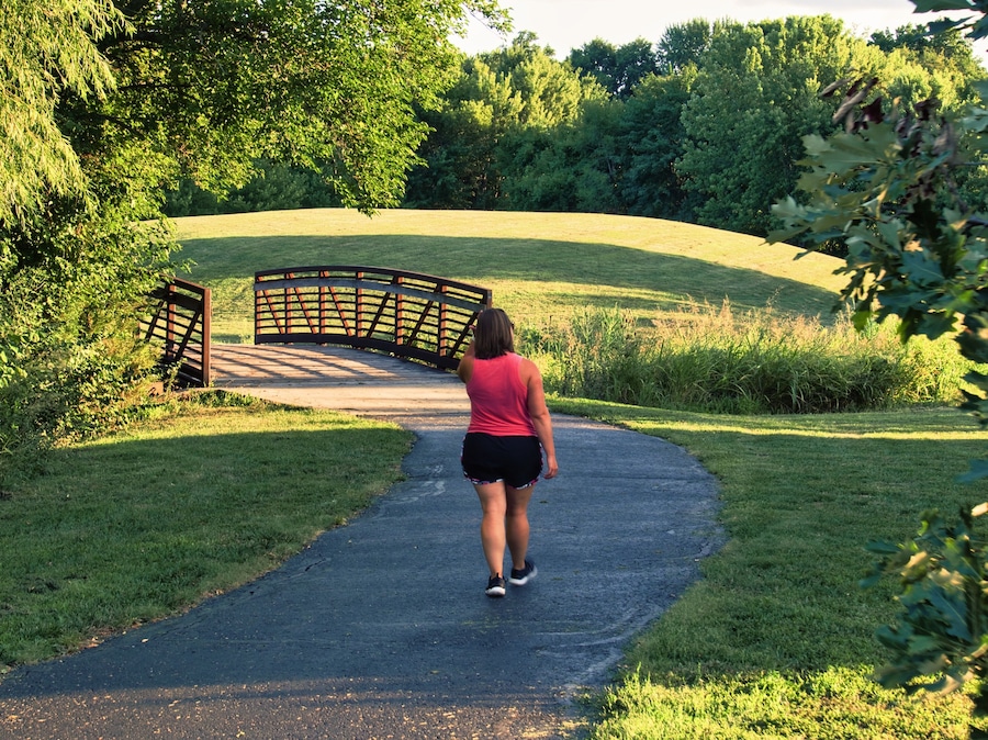 Uma mulher caminhando por um caminho pavimentado em um parque, com uma ponte de madeira ao fundo.