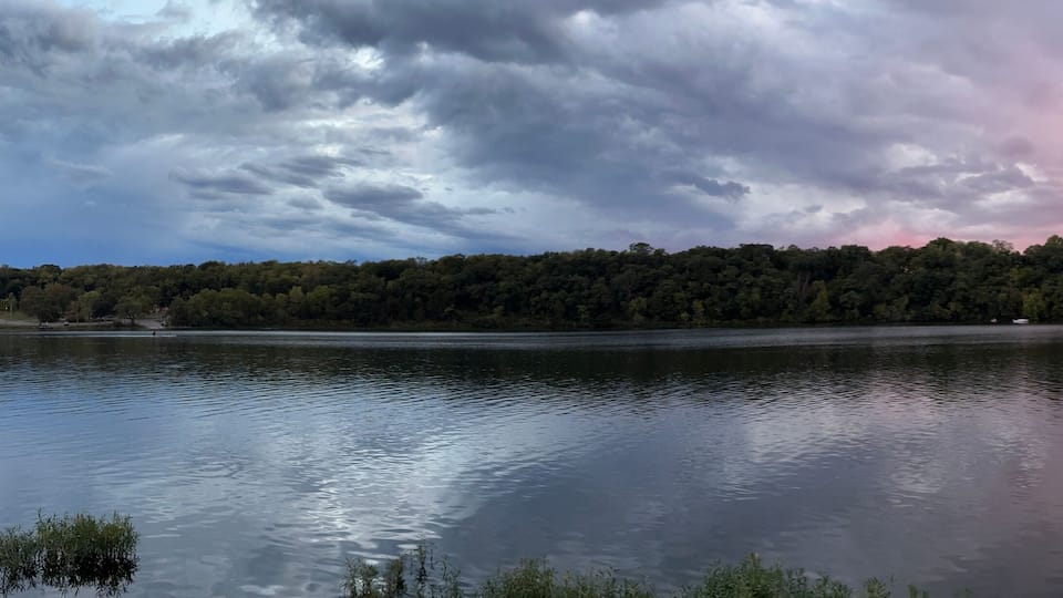 Un lago tranquilo con una ribera arbolada y un cielo espectacular con tonos rosados y morados.