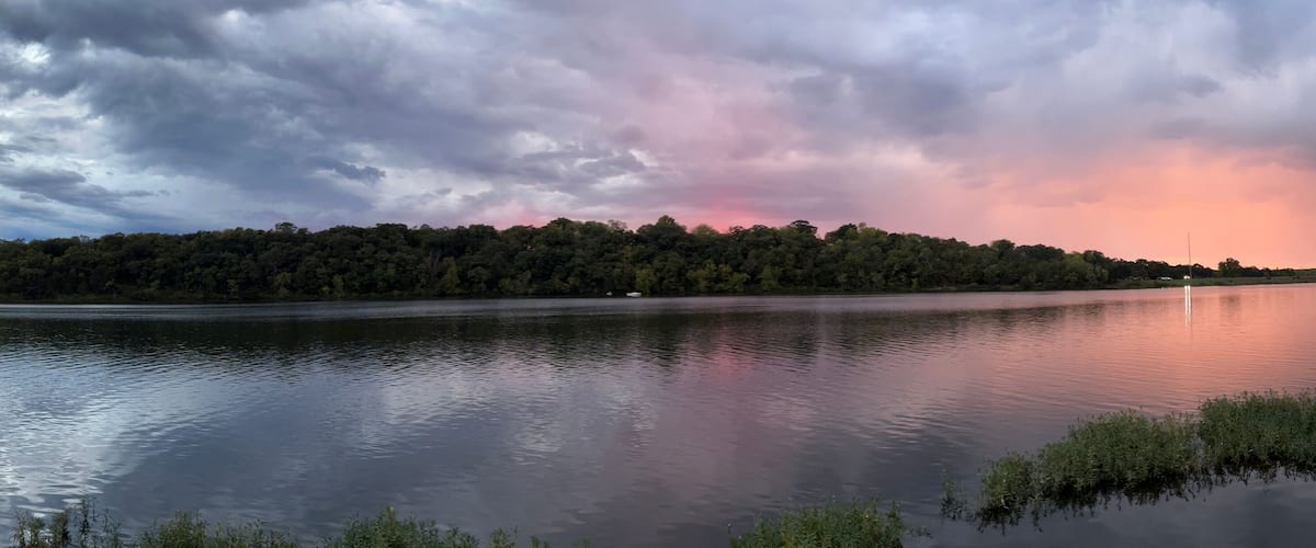 A calm lake with a forested shoreline and a dramatic sky with pink and purple hues.