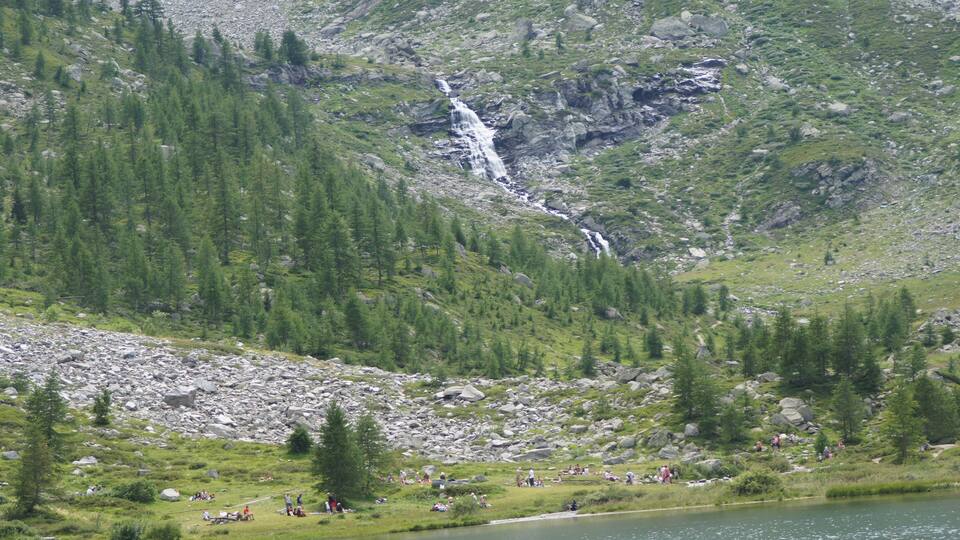 A waterfall cascading down a rocky hillside, surrounded by dense green forest and a group of people enjoying the outdoor setting by a lake.