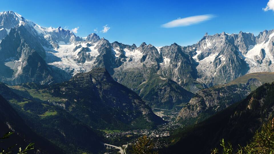 A mountainous landscape with snow-capped peaks, dense forests, and a valley with a small settlement.