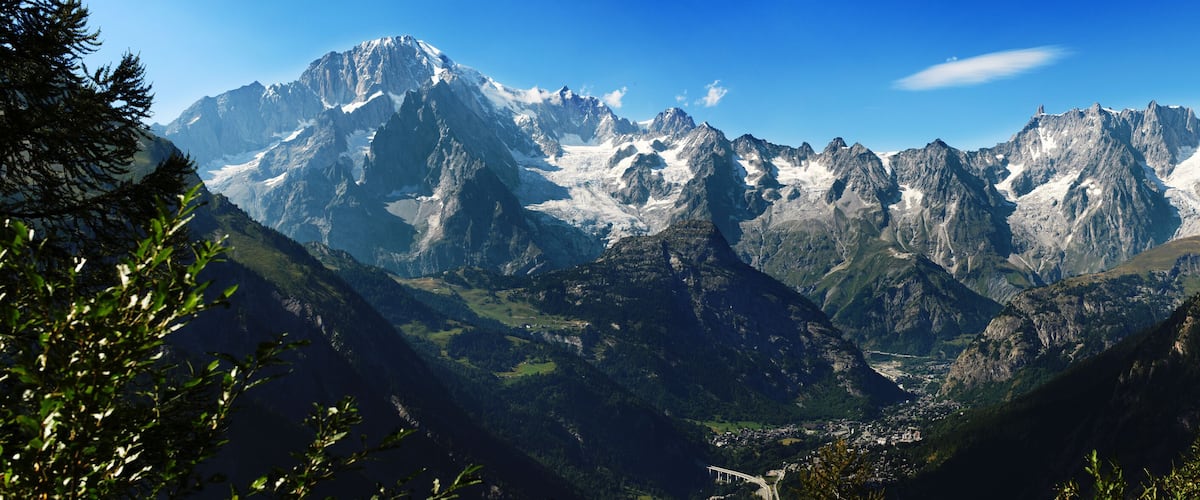 A mountainous landscape with snow-capped peaks, dense forests, and a valley with a small settlement.