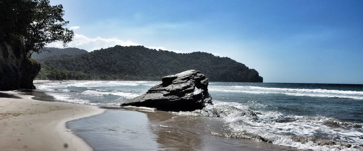 A beach with a large rock in the foreground, waves crashing onto the shore, and a forested hill in the background.