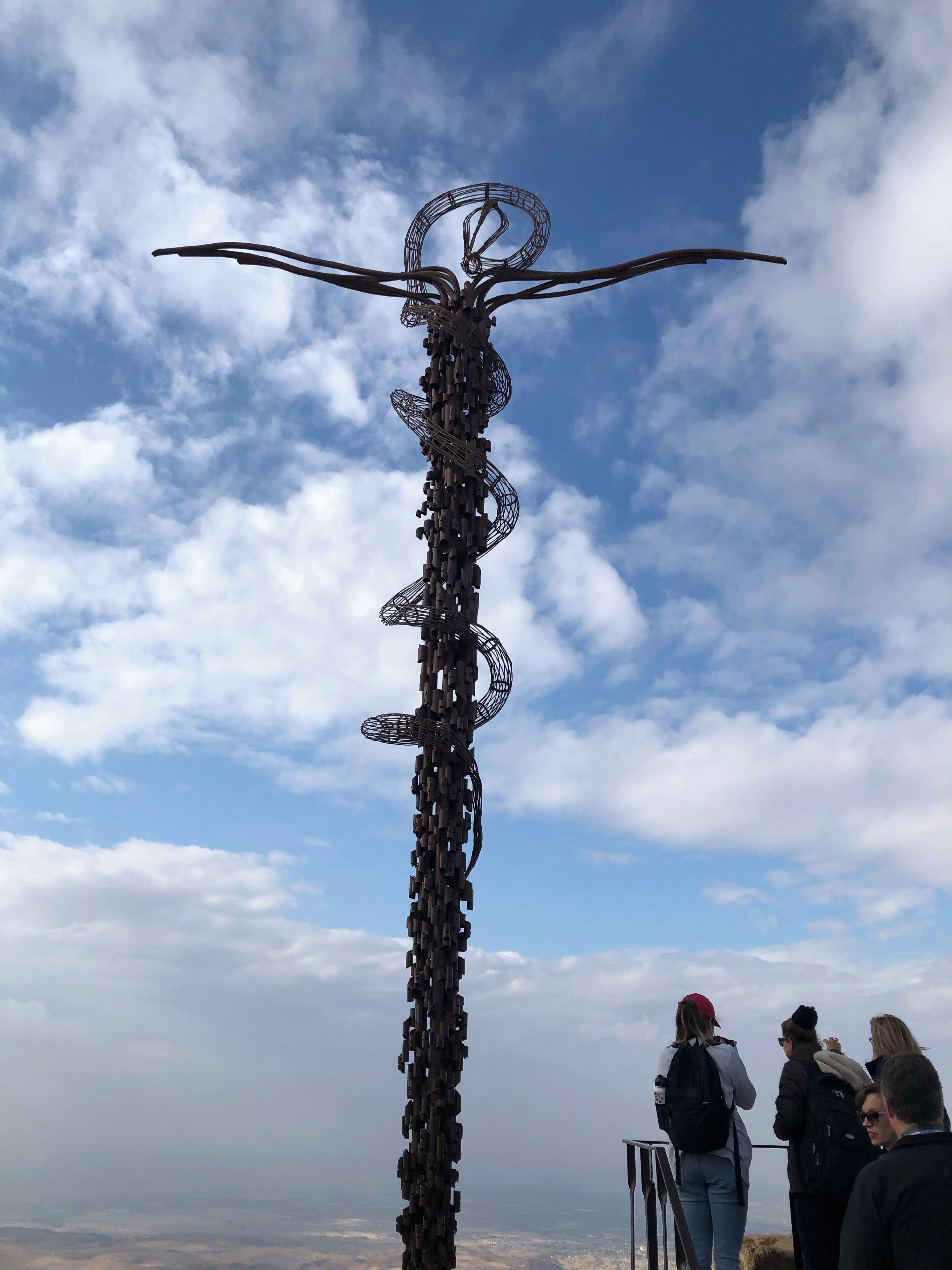Una alta escultura de metal con una parte superior circular y múltiples brazos, rodeada por un paisaje montañoso y un cielo parcialmente nublado.