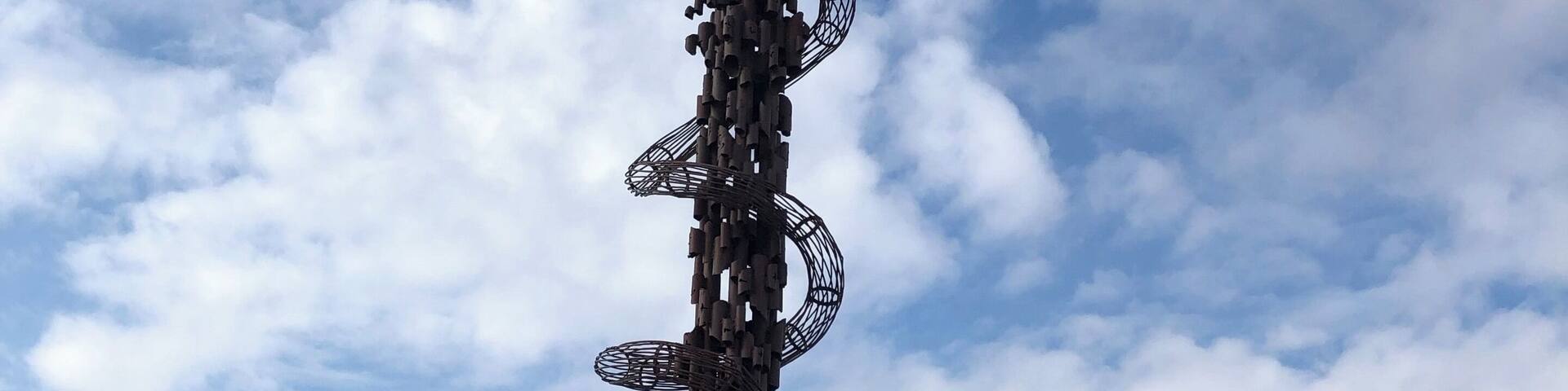 A tall metal sculpture with a circular top and multiple arms, surrounded by a mountainous landscape and a partly cloudy sky.