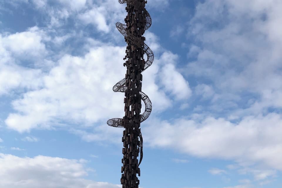 A tall metal sculpture with a circular top and multiple arms, surrounded by a mountainous landscape and a partly cloudy sky.