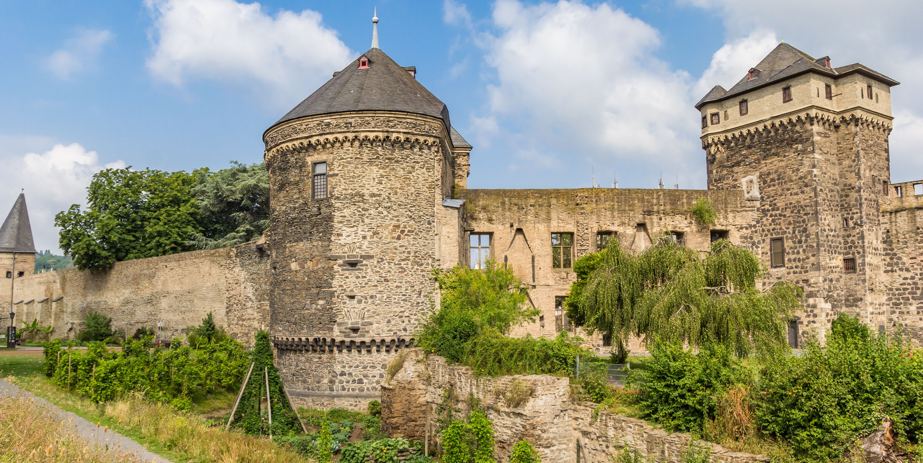 Un château historique en pierre, doté d’une tour ronde, entouré d’une végétation luxuriante et surplombé d’un ciel dégagé.
