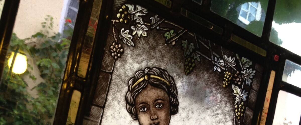 A stained glass window with a woman holding a chalice, surrounded by foliage and a cityscape.