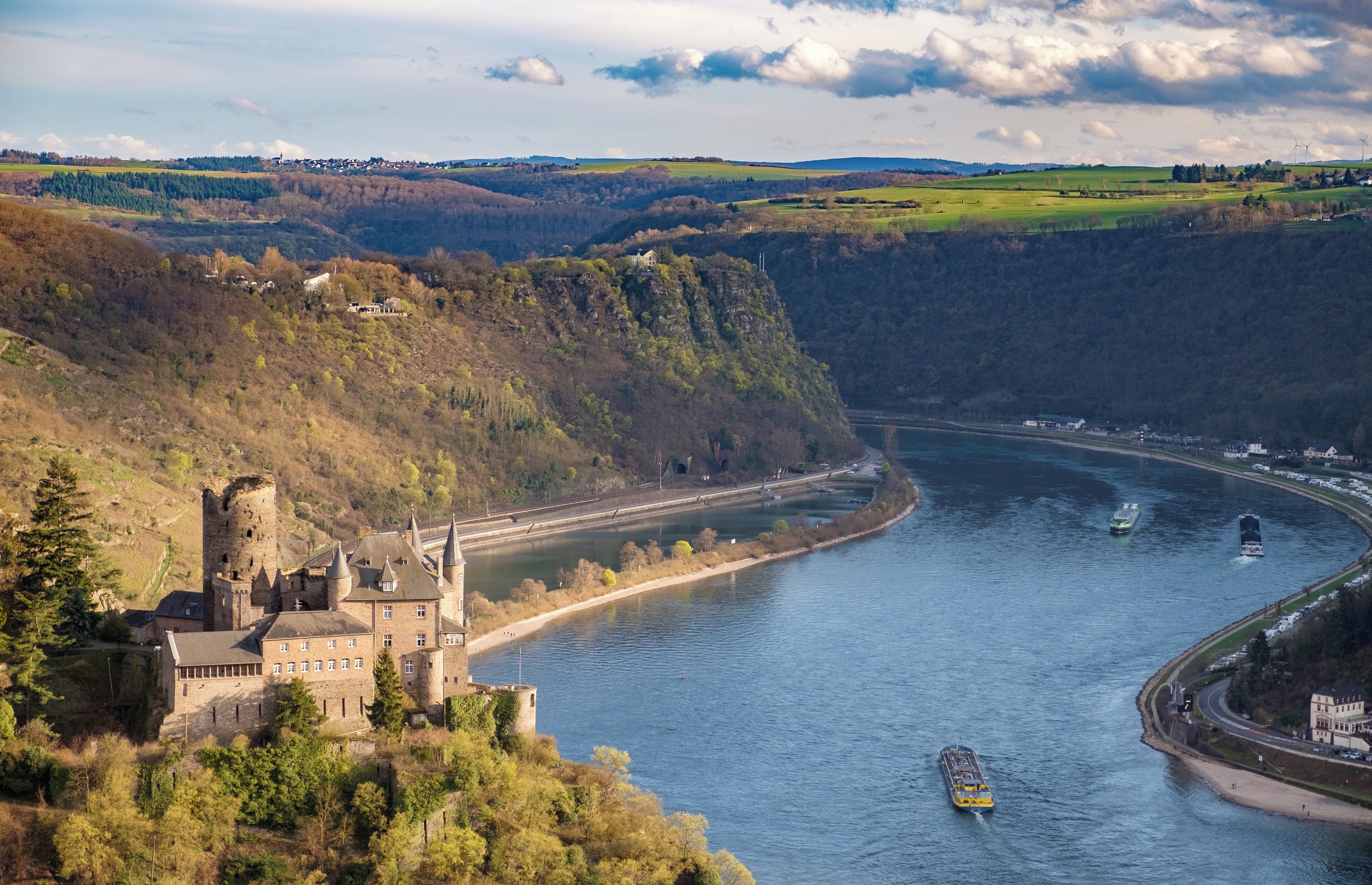 A castle by a river with boats and wind turbines in the background.