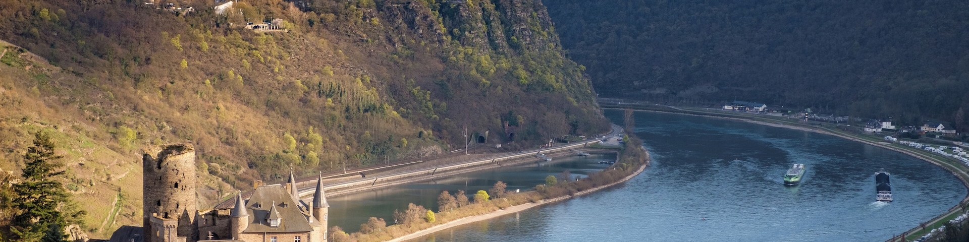 Un castillo a orillas de un río, con barcos y aerogeneradores al fondo.