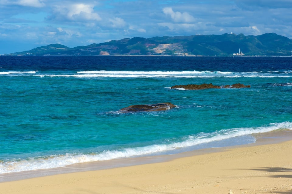A beach with turquoise waters, a sandy shore, and a mountain range in the background.