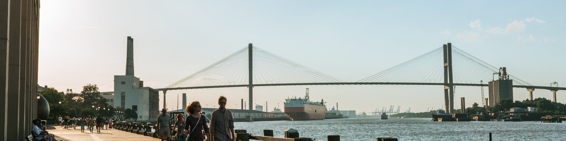 Un paseo peatonal junto a un río, con un puente grande al fondo.
