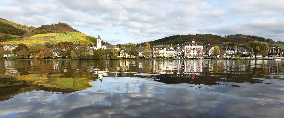 A calm lake with a town on the opposite shore, featuring a church steeple and a hill with vineyards.