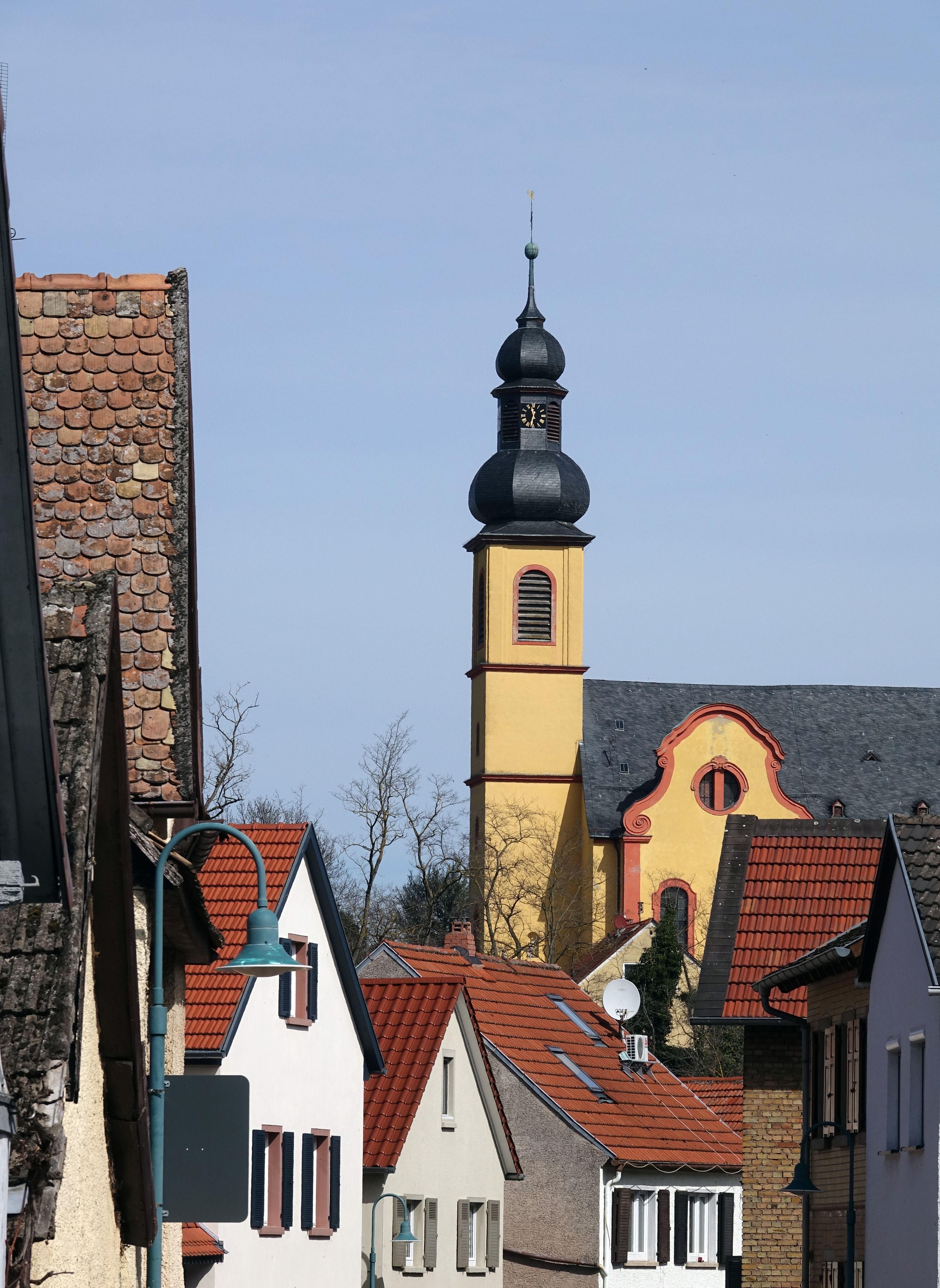 A church with a yellow tower and a black spire, surrounded by traditional European houses with red roofs.