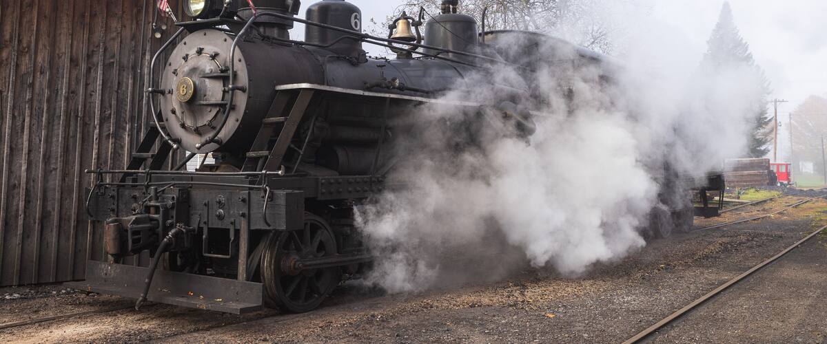 A steam locomotive emitting smoke, with a wooden building and a red train car in the background.